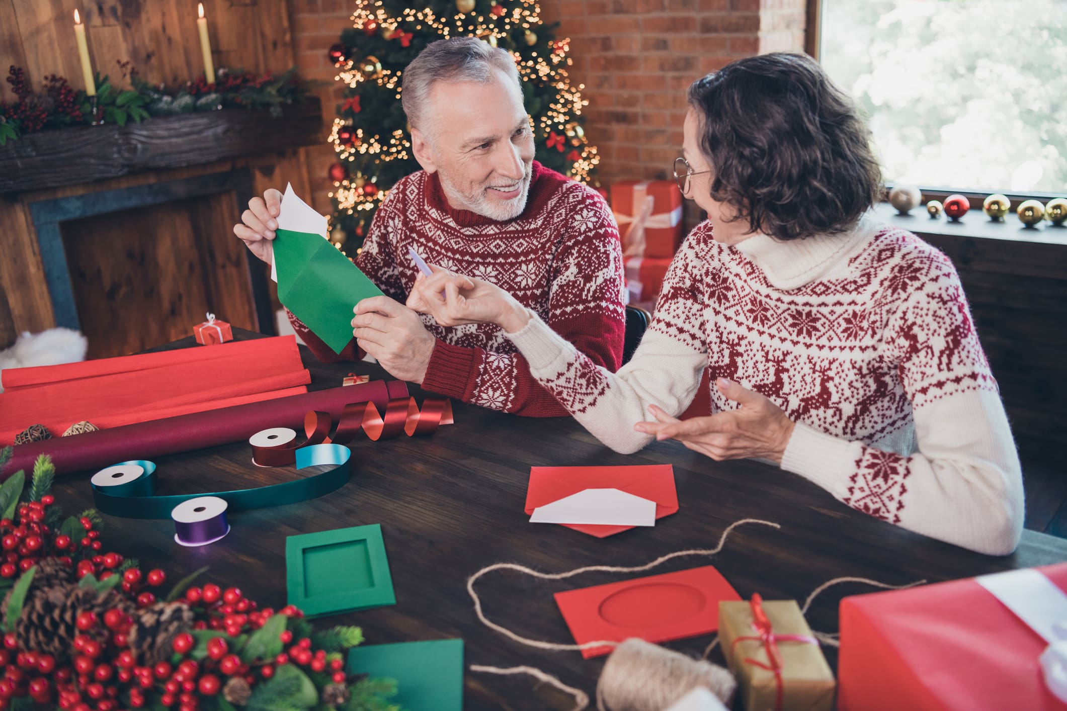 Photo of happy positive couple talking, grandma and grandpa while packing envelop letter wearing ornament sweaters home indoor
